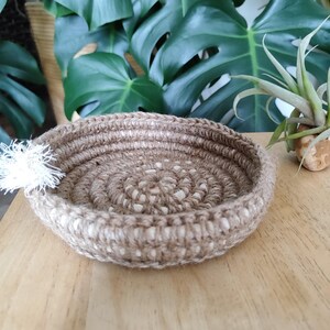 May include: A round, woven jute basket with a natural brown and white color scheme. The basket has a spiral design and a small white tassel. The basket is on a wooden surface, with green leaves in the background.