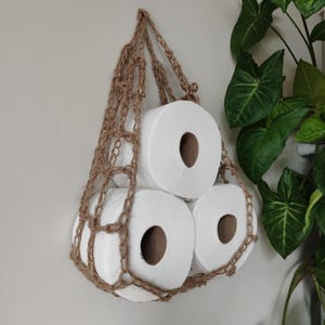 May include: Three rolls of white toilet paper are held in a brown crocheted hanging storage basket.