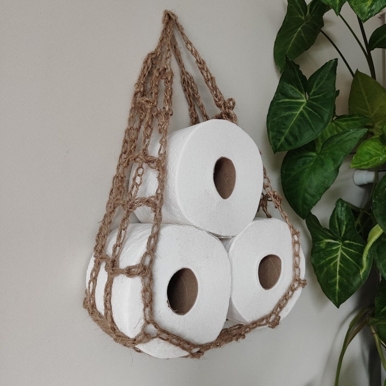May include: Three rolls of white toilet paper are held in a brown crocheted hanging storage basket.