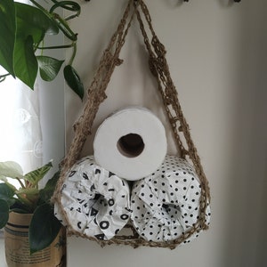 May include: Three rolls of toilet paper hanging from a macrame wall hanging. The rolls are white with black polka dots and one is plain white.