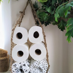 May include: A hanging toilet paper holder made of natural jute rope with five rolls of toilet paper. The rolls are white and have different patterns, including polka dots and geometric shapes.