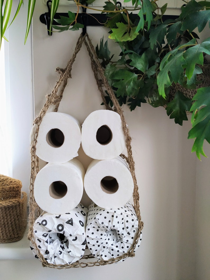 May include: A hanging toilet paper holder made of natural jute rope with five rolls of toilet paper. The rolls are white and have different patterns, including polka dots and geometric shapes.