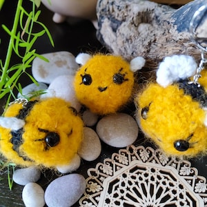 May include: Three crocheted bee ornaments with black eyes and smiling faces. The bees are yellow and black and have white wings. The bees are sitting on a wooden surface with a decorative pattern.