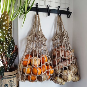 May include: Two brown crocheted string bags hanging from hooks on a white wall. One bag is filled with oranges and the other with onions.