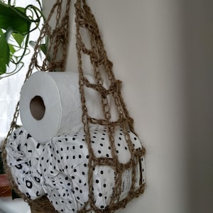 May include: A brown crocheted hanging storage bag with a roll of white toilet paper and a roll of patterned toilet paper inside.