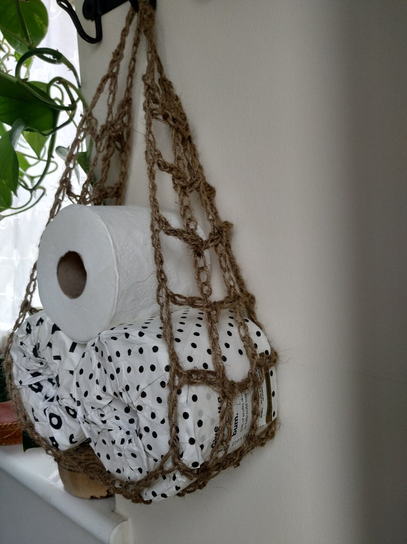 May include: A brown crocheted hanging storage bag with a roll of white toilet paper and a roll of patterned toilet paper inside.