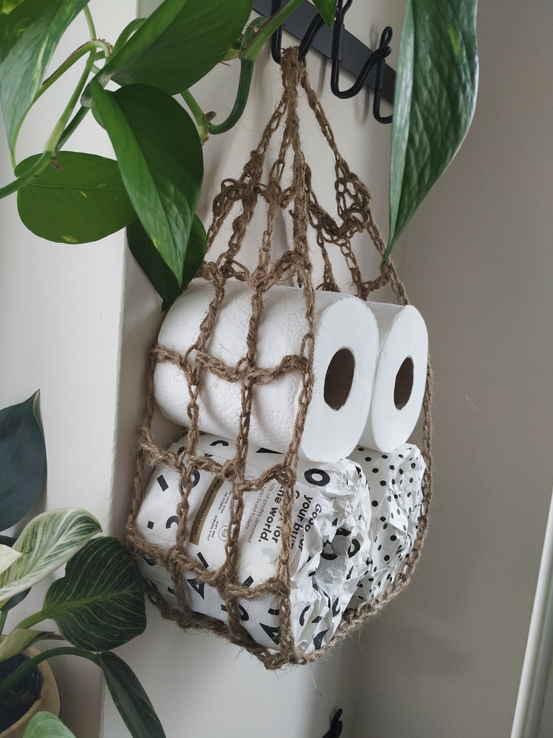 May include: A brown crocheted hanging storage net with two rolls of toilet paper inside. The toilet paper is wrapped in white paper with black polka dots and a white paper with black text that reads "GOOD. your bu or ne world. 10% of profits donated to help build toilets."