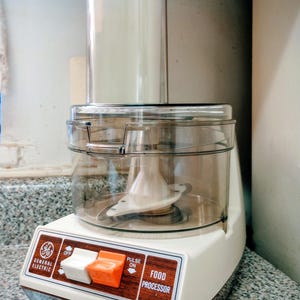 Peut inclure: Un robot culinaire General Electric vintage. L'appareil a une base blanche avec une garniture marron et un bol en plastique transparent. Il comporte des boutons blancs et orange et les mots "Food Processor".