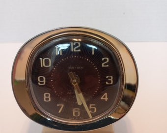 May include: A silver and white Baby Ben alarm clock with black numbers and hands. The clock face is dark brown and says "Baby Ben" and "Westclox" in white lettering. The clock is made in the USA.