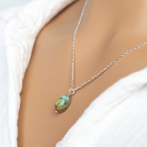 May include: A silver chain necklace with a small, oval-shaped turquoise pendant. The pendant has a mix of blue, green, and brown colors. The necklace is worn against a white shirt.