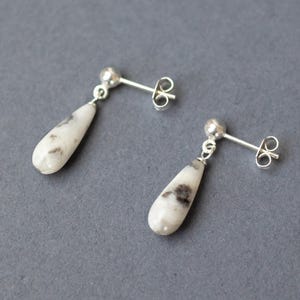 May include: A pair of silver drop earrings with teardrop-shaped white and gray speckled stones. The earrings have a post and butterfly backing. The background is a solid gray.