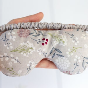 May include: A gray eye mask with a floral pattern featuring white flowers, green leaves, and pink and red berries. The mask has an elastic band for a comfortable fit.