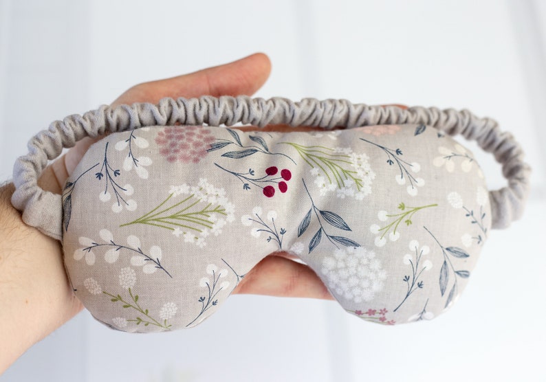 May include: A gray eye mask with a floral pattern featuring white flowers, green leaves, and pink and red berries. The mask has an elastic band for a comfortable fit.