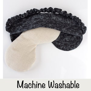 May include: A black and white eye mask with a white, u-shaped pillow inside. The eye mask is made of a soft, textured fabric and has a ruffled edge. The text "Machine Washable" is printed below the image.