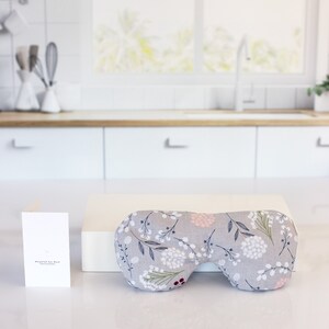 May include: A gray eye mask with a floral pattern in shades of white, pink, and green. The eye mask is on a white surface with a white card that says "Wrapped Eye Mask" in black text.