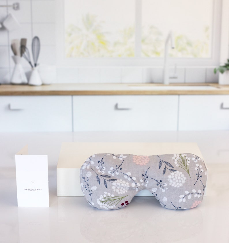 May include: A gray eye mask with a floral pattern in shades of white, pink, and green. The eye mask is on a white surface with a white card that says "Wrapped Eye Mask" in black text.