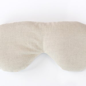 May include: A beige linen eye pillow with a curved shape. The pillow is designed to fit over the eyes and is filled with a soft material.