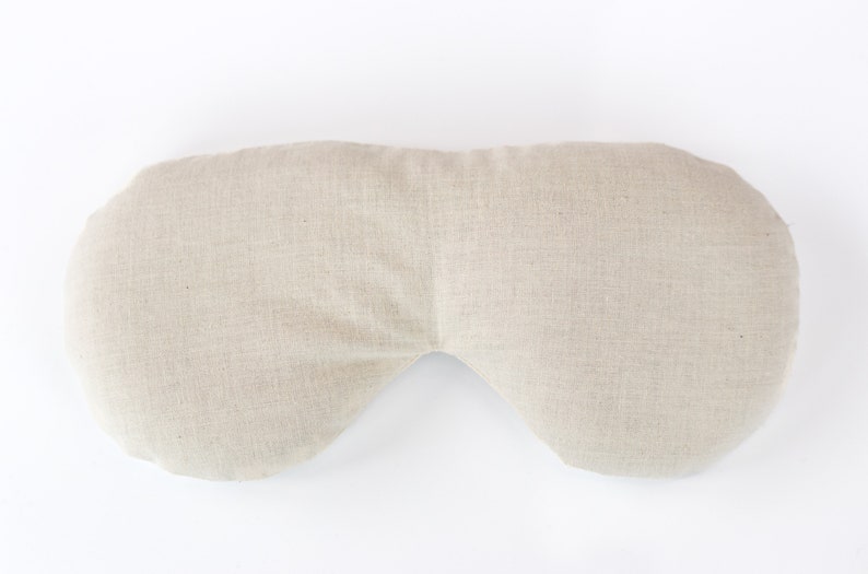 May include: A beige linen eye pillow with a curved shape. The pillow is designed to fit over the eyes and is filled with a soft material.