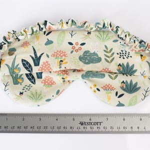 May include: A white sleep mask with a colorful pattern of mushrooms, flowers, and birds. The mask is made of soft fabric and has an elastic band for a comfortable fit.