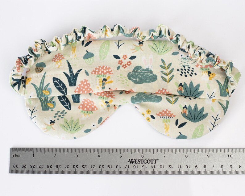 May include: A white sleep mask with a colorful pattern of mushrooms, flowers, and birds. The mask is made of soft fabric and has an elastic band for a comfortable fit.