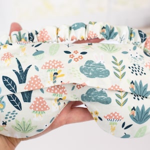 May include: A white sleep mask with a colorful pattern of mushrooms, flowers, and leaves. The pattern is reminiscent of a whimsical forest scene.