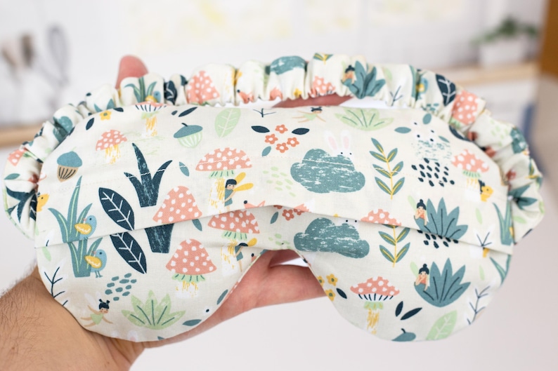 May include: A white sleep mask with a colorful pattern of mushrooms, flowers, and leaves. The pattern is reminiscent of a whimsical forest scene.