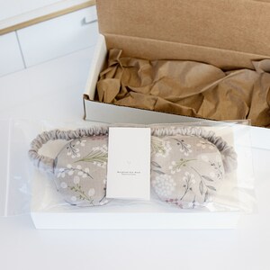 May include: A gray eye mask with a white floral pattern and a white tag that reads "Weighted for Rest" is packaged in a clear plastic bag. The bag is sitting on top of a white box.