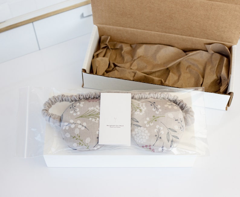May include: A gray eye mask with a white floral pattern and a white tag that reads "Weighted for Rest" is packaged in a clear plastic bag. The bag is sitting on top of a white box.