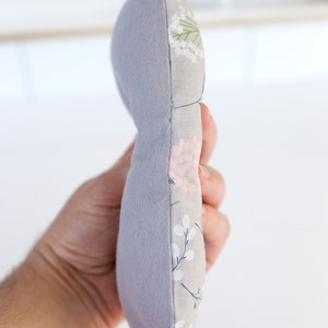 May include: A gray and white floral fabric pillow with a soft, plush texture. The pillow is shaped like a crescent moon and is sewn together with a visible seam.