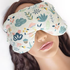 May include: A white sleep mask with a whimsical forest print featuring mushrooms, flowers, and woodland creatures. The mask is made of soft fabric and has a contoured shape for comfortable wear.