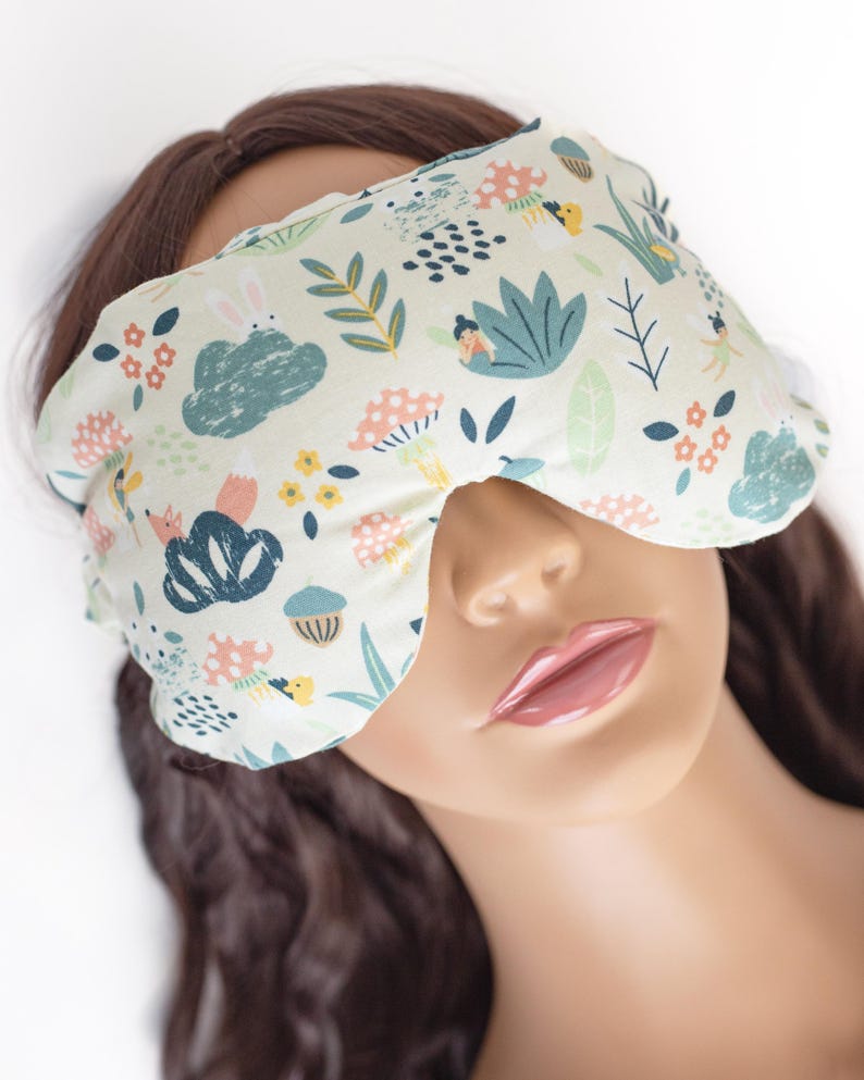 May include: A white sleep mask with a whimsical forest print featuring mushrooms, flowers, and woodland creatures. The mask is made of soft fabric and has a contoured shape for comfortable wear.