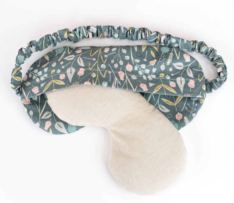 May include: A teal blue sleep mask with a floral pattern and a light beige fabric lining. The mask has an elastic band for a comfortable fit.