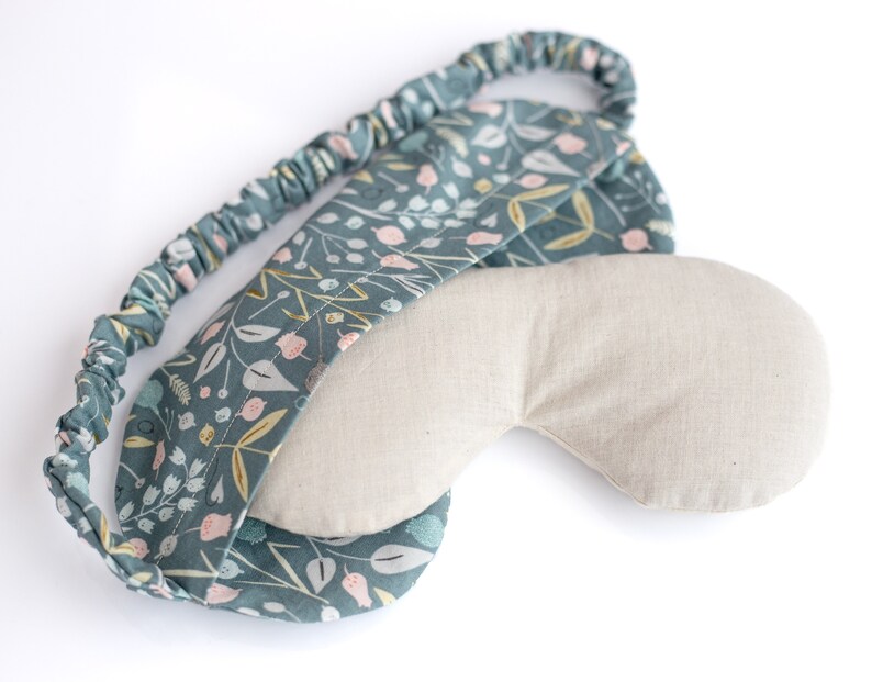 May include: A teal blue sleep mask with a floral pattern and a light beige fabric lining. The mask has an elastic band for a comfortable fit.