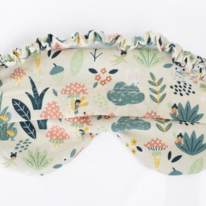 May include: A light beige sleep mask with a whimsical forest print featuring fairies, mushrooms, birds, and leaves. The mask has an elastic band for a comfortable fit.