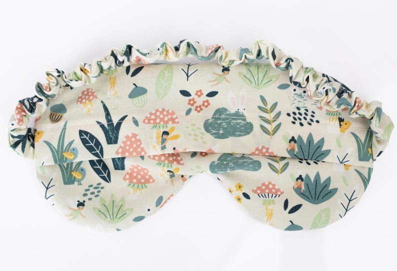 May include: A light beige sleep mask with a whimsical forest print featuring fairies, mushrooms, birds, and leaves. The mask has an elastic band for a comfortable fit.