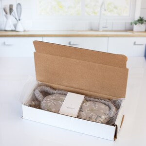 May include: A white cardboard box with a floral eye mask inside. The box is open and the eye mask is visible. The eye mask is made of a soft fabric with a floral print. The text on the box reads "Whisper the Moon".