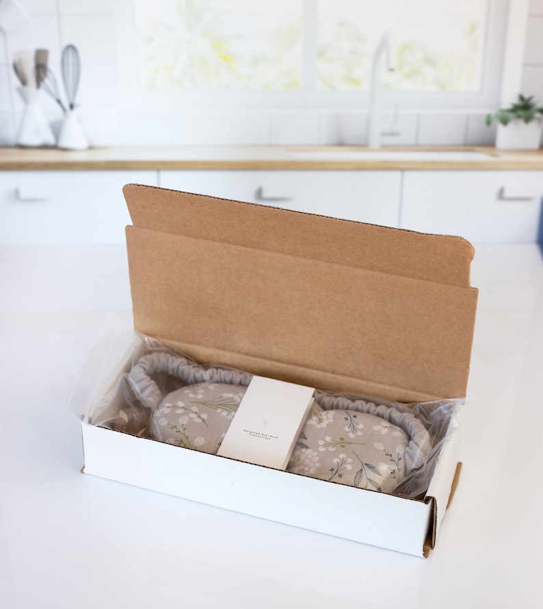 May include: A white cardboard box with a floral eye mask inside. The box is open and the eye mask is visible. The eye mask is made of a soft fabric with a floral print. The text on the box reads "Whisper the Moon".