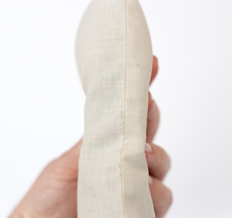 May include: A close-up of a cream-colored fabric pouch with a seam down the center. The pouch is held in a hand.