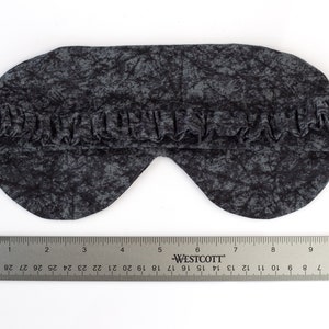 May include: A black sleep mask with a gray and black abstract pattern. The mask is made of soft fabric and has an elastic band for a comfortable fit.