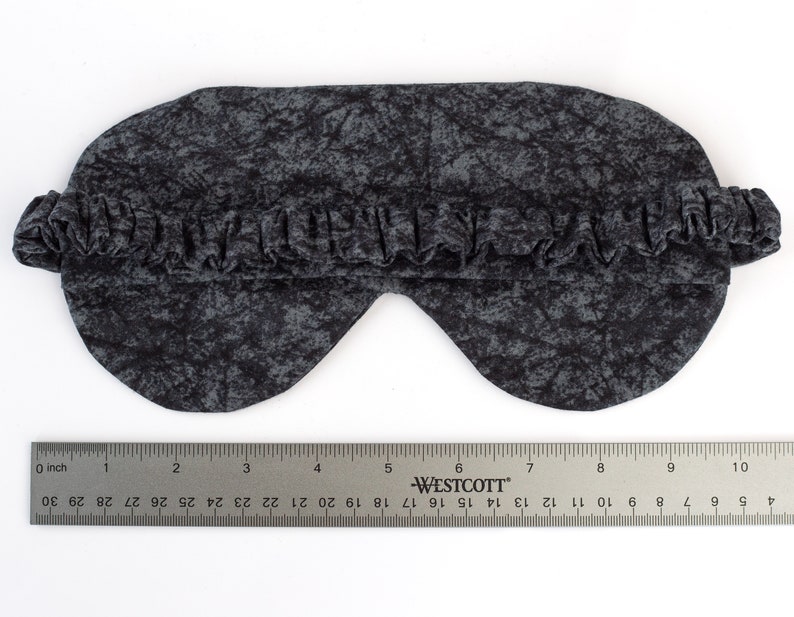 May include: A black sleep mask with a gray and black abstract pattern. The mask is made of soft fabric and has an elastic band for a comfortable fit.