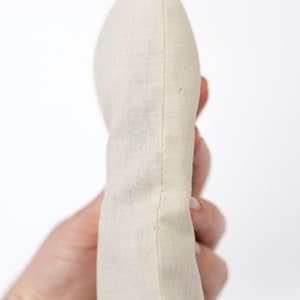 May include: A close-up of a cream-colored fabric pouch with a seam down the center. The pouch is held in a hand.