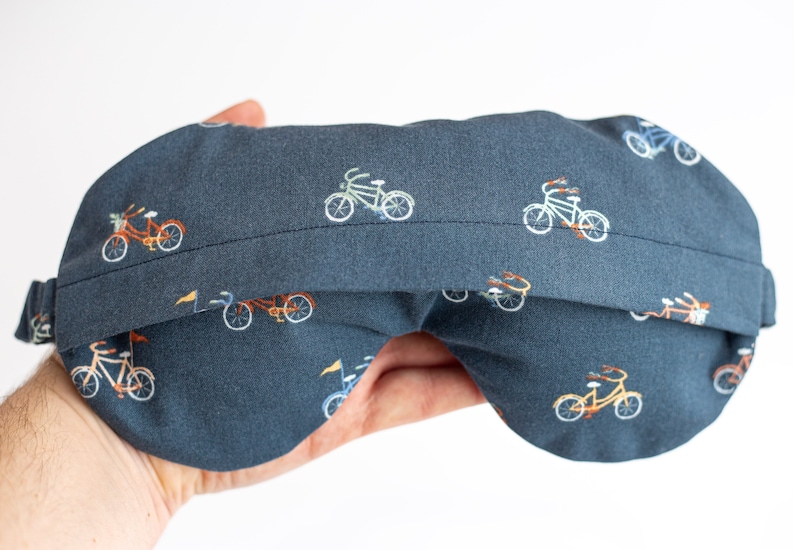 以下が含まれることがあります： カラフルな自転車が繰り返し描かれた濃い青色のアイマスク。柔らかな生地で作られており、快適な睡眠のための輪郭のある形状をしています。