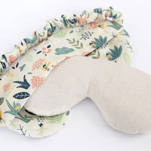 May include: A beige eye mask with a floral print on one side. The print features a light green, pink, and yellow color scheme with small illustrations of flowers, leaves, and birds.