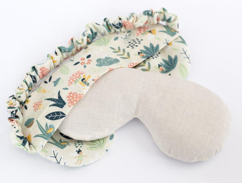 May include: A beige eye mask with a floral print on one side. The print features a light green, pink, and yellow color scheme with small illustrations of flowers, leaves, and birds.