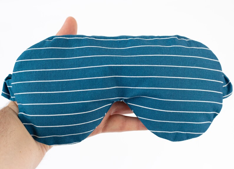 Puede incluir: M&aacute;scara para dormir a rayas blancas y azul verdoso. La m&aacute;scara est&aacute; hecha de tela y tiene un patr&oacute;n de rayas horizontales. La m&aacute;scara tiene una forma curva para adaptarse a los ojos y tiene correas de tela a cada lado.