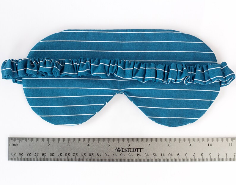 Puede incluir: M&aacute;scara para dormir a rayas blancas y azul verdoso con una banda el&aacute;stica con volantes. La m&aacute;scara mide aproximadamente 20 cm de largo, con una regla en la imagen para la escala. La m&aacute;scara est&aacute; dise&ntilde;ada para bloquear la luz.