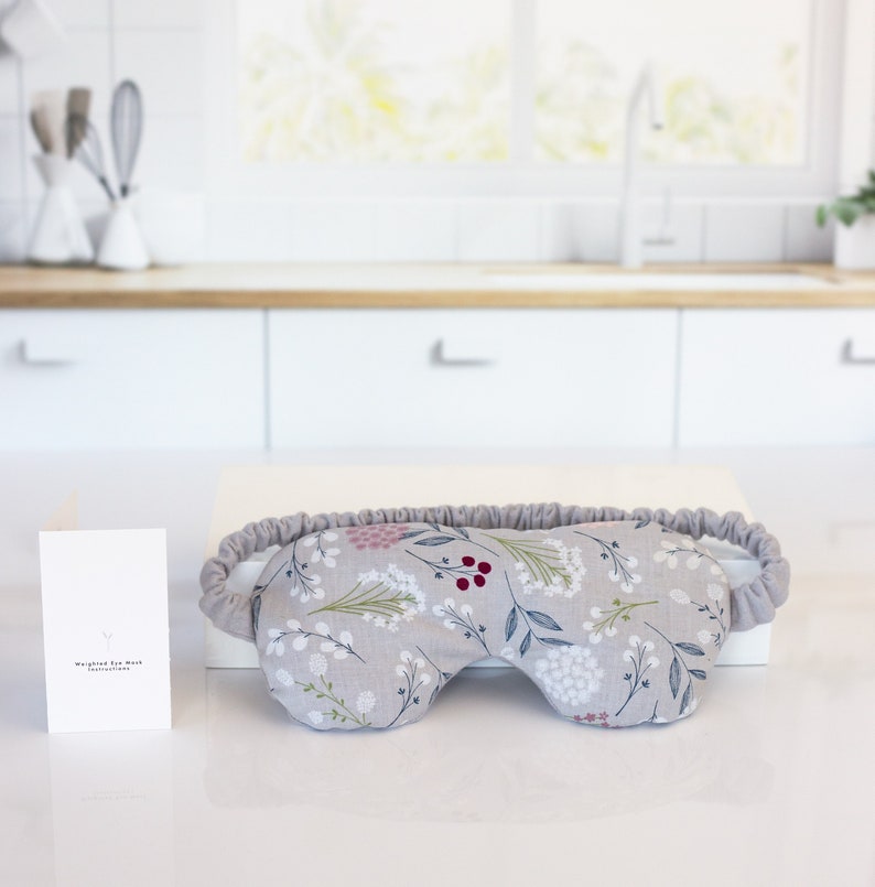 May include: A gray eye mask with a floral pattern featuring white, pink, green, and red flowers. The eye mask is made of soft fabric and has an elastic band.
