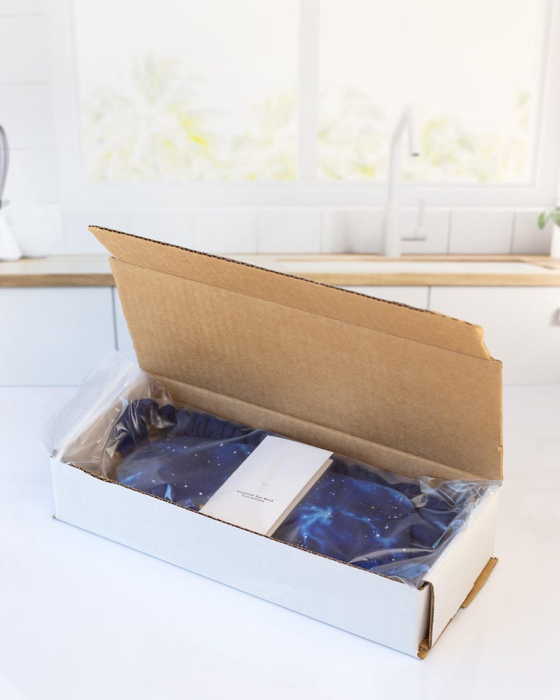 May include: A blue sleep mask with a galaxy print inside a white cardboard box. The box is open and the sleep mask is visible.