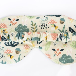May include: A cream-colored sleep mask with a whimsical forest print featuring mushrooms, leaves, and bunnies. The mask has an elastic band for a comfortable fit.
