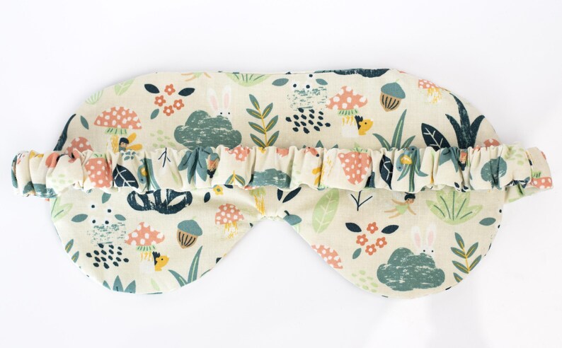 May include: A cream-colored sleep mask with a whimsical forest print featuring mushrooms, leaves, and bunnies. The mask has an elastic band for a comfortable fit.